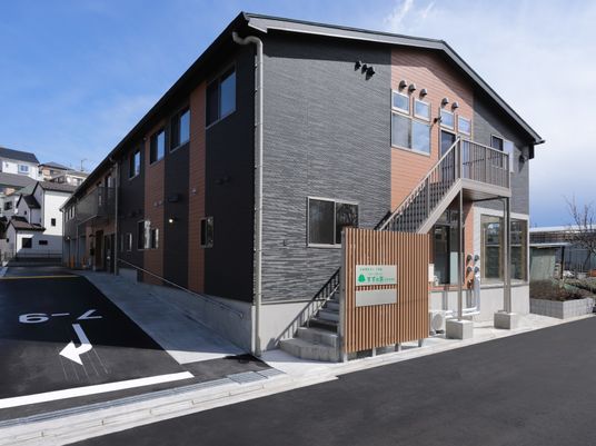 2階建ての建物。ベージュとグレーで屋外階段やベランダが付き、玄関には手すり付きのスロープが付いている。
