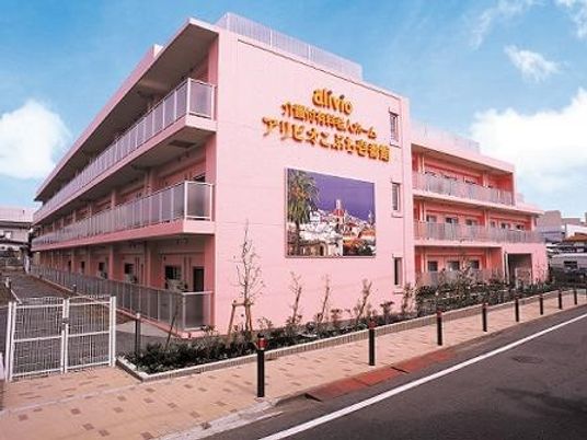 ピンク色の３階建ての建物。周辺には木が植えられ、駐車場も完備。