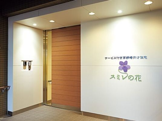 白い壁には住宅名が書かれ、すみれの花の絵が描かれている。ライトアップされた玄関には木製のドアが付いている。