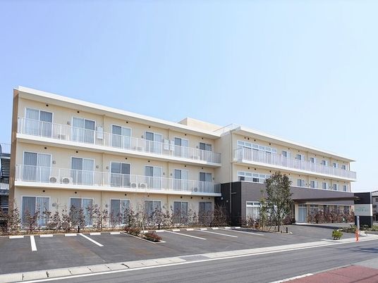 駐車場付きの3階建ての建物は白く、玄関には車寄せが付き、建物周辺には緑が植えられている。