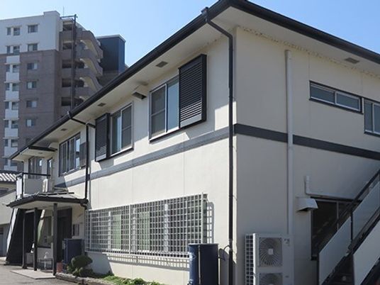 ダークグレーの2階建ての建物はモダンなデザイン。周辺には緑が多く、玄関前には手すり付きの階段が付いている。