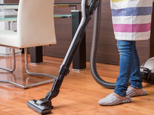 ボーダー柄のエプロンを付けた女性スタッフがフローリングの部屋に掃除機をかけている様子。