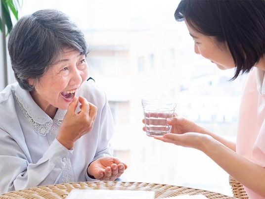 薬を飲もうとしている高齢女性のそばには水の入ったコップを持った女性スタッフがいてほほ笑んでいる。