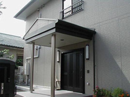 グレー系の外観で２階建ての建物は玄関に屋根があり、草木が周りに植えられている。