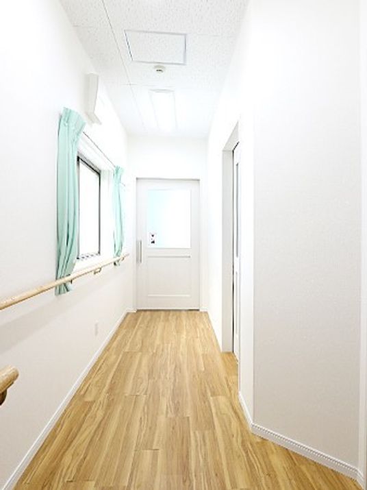 木目調の床と手すり付きの壁が写った施設内の廊下の様子。左側に窓、右側に個室のドア