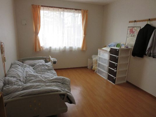 寝具付きのベッドが置かれたフローリングの部屋。プラスチックケースが置かれ、レースカーテンとカーテンが付いた大きな窓がある。