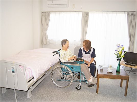 車いすの乗った高齢者と施設スタッフが会話を交わしている様子。施設内の居室の一場面