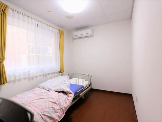 小さなワンルームタイプの部屋。エアコンと窓があり、窓辺にベッドが置いてある