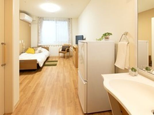介護用ベッドと冷蔵庫、椅子、テーブルなどが置かれている施設内の居室の様子