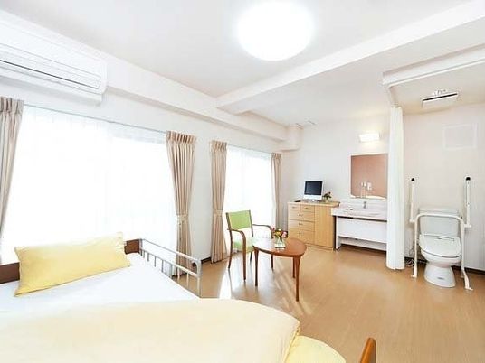 広めのワンルームにトイレや洗面台などがついている様子。手前にベッドが置いてある