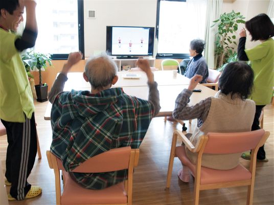 テレビを見ながら体操している高齢者と施設のスタッフの様子。広めのテーブルが置いてあるスペース