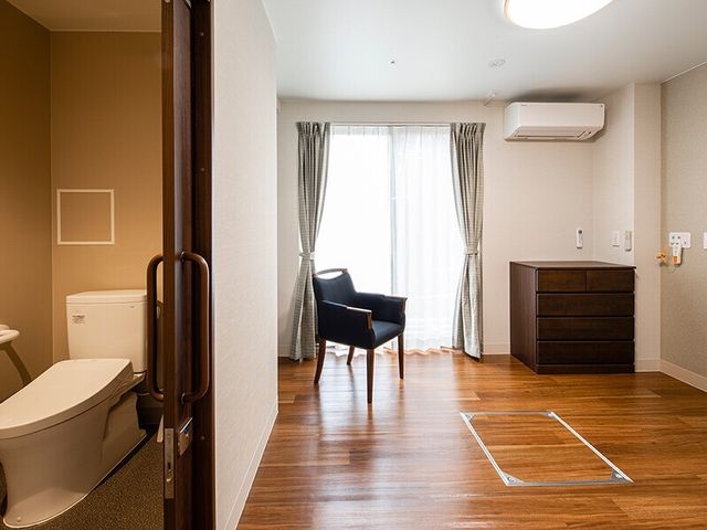 フローリングの部屋にはいすやタンスが置かれ、緊急通報装置が付き、引き戸が付いたトイレもある。 アクセス良好で便利な立地がなじみの関係や今まで通りの暮らしをキープできるようサポート！医療連携もあり、最期まで安心して暮らせる体制です。