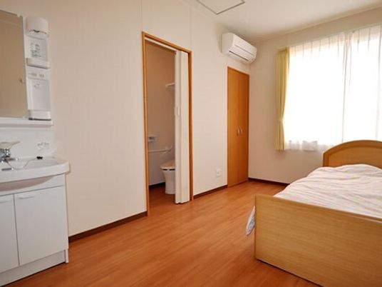 トイレと洗面所が付いたフローリングの部屋には大きな窓があり、寝具付きのベッドやエアコンも設置されている。