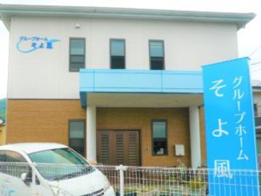 白とベージュの2階建ての建物にはライトブルーの屋根が付いた玄関があり、玄関の前にはブルーの看板も建っている。