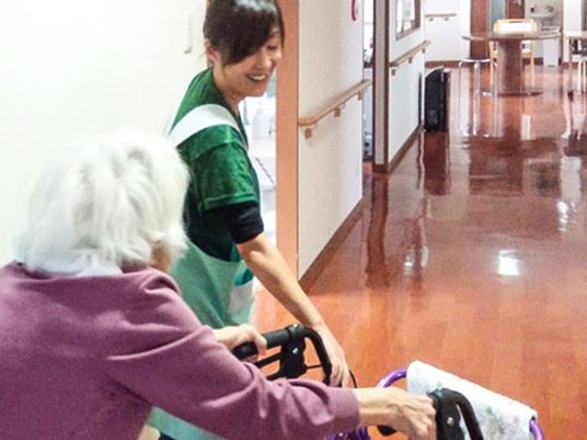 まっすぐな廊下をシルバーカーを使用して歩いている高齢女性。エプロンを付けた女性スタッフが付き添っている。