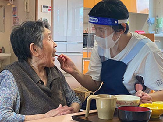 フェイスガードやマスクを付けた女性スタッフが高齢女性の食事介助を行っている様子。