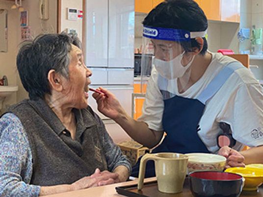 フェイスガードやマスクを付けた女性スタッフが高齢女性の食事介助を行っている様子。