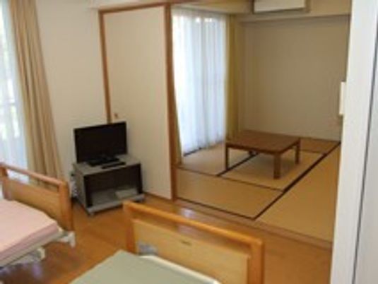 テレビや介護ベッドが2台置かれたフローリングの部屋が手前に、奥には畳敷きの和室がある。