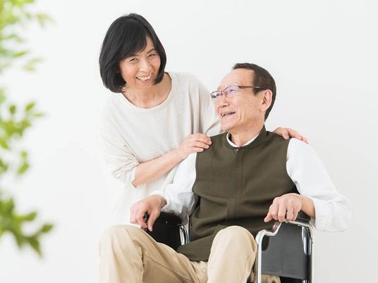 白いゆったりとした服を着た高齢女性と車いすの高齢男性が笑顔で話している。