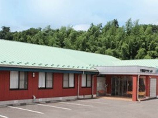 ライトグリーンの屋根が付いている平屋建ての建物はエンジ系の外壁で緑が多く周りにある。