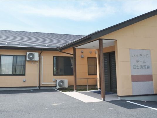 ベージュの外壁の平屋建ての建物は日当たりのいい場所に建っている。玄関には屋根もある。