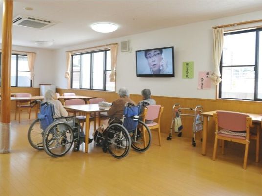 食堂のテーブルを囲んでテレビを見ている高齢女性たち。車いすに座っている人もいる。