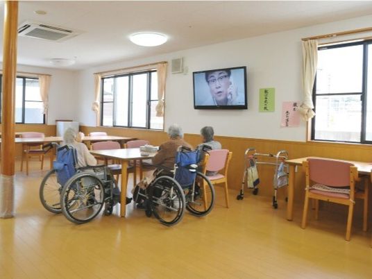 食堂のテーブルを囲んでテレビを見ている高齢女性たち。車いすに座っている人もいる。