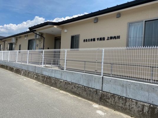 住宅名が書かれたベージュの外壁の平屋建ての建物。玄関には屋根が付いている。