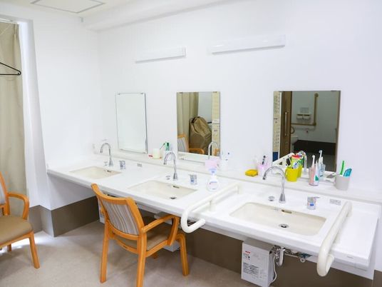 洗面所が３つ並んでいて、それぞれに鏡が付いている。中央の洗面所にはいすもセットされている。