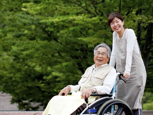 車いすに乗った男性入居者と散歩を楽しむ女性スタッフ。自然あふれる背景。