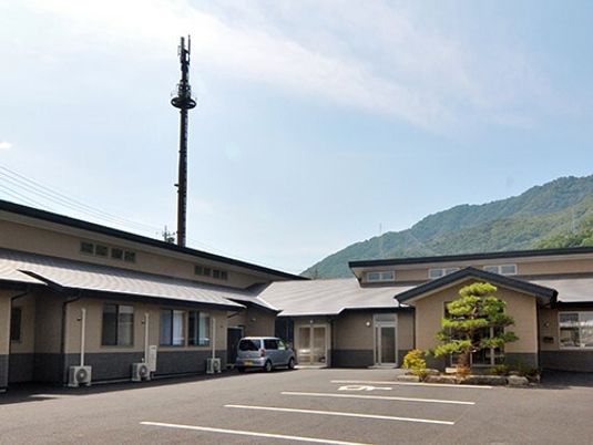 後方には緑あふれる山があり、前方には広い駐車場がある平屋建ての建物。車も止まっている。
