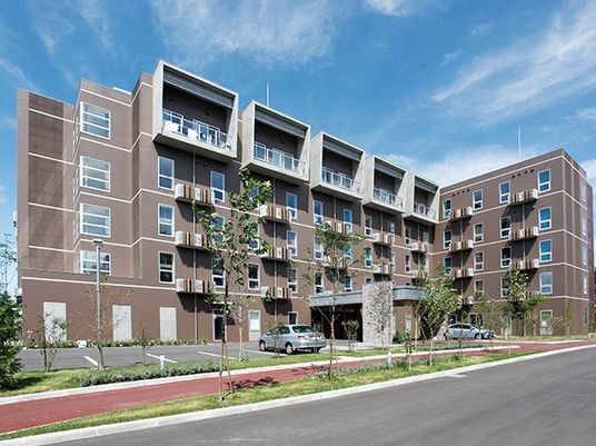 建物外観、昼間。５階建ての建物の前には駐車場、そして道路わきには遊歩道がある。青空が広がり明るい雰囲気。