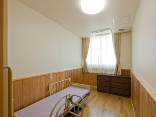 ベッドが置かれたフローリングの部屋にはタンスやエアコンがあり、レースカーテンとカーテンが付いた窓がある。