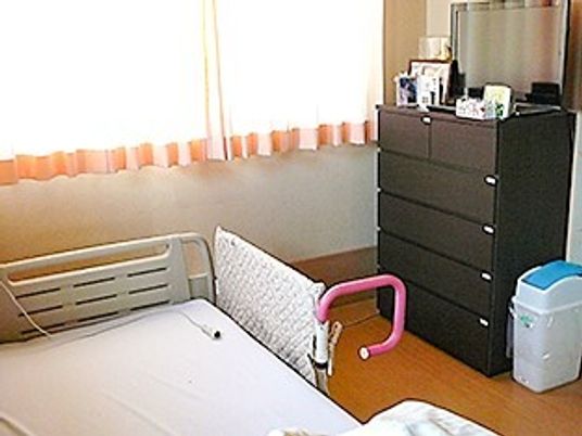 寝具付きのベッドが置かれ、レースカーテンとカーテンが付いた窓があり、タンスも置かれている。