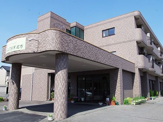 グレー系の３階建ての建物は各フロアにベランダがあり、建物周辺には植物が置かれ、車寄せも付いている。