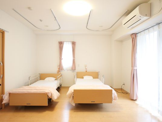 二人用の居室にベッドが二つ並び、壁にはエアコンが設置されている。窓が大きく日当たりがいい部屋。
