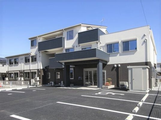 広い駐車場が付いた大きな建物は3階建て。白とグレーのモノトーンで落ち着いた雰囲気。玄関には車寄せと自動ドアが設置されている。
