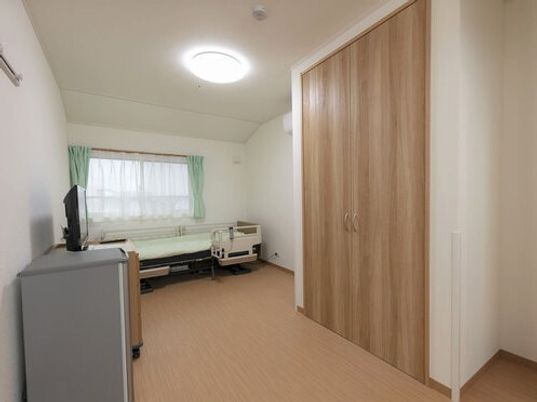 大きなクローゼットやタンス、テレビ、介護ベッドが設置された部屋には大きな窓も付き、カーテンとレースカーテンが付いている。