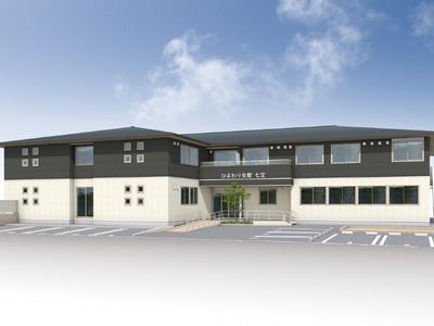 モノトーンの2階建ての建物。ホーム名が書かれた屋根と手すり付きのスロープが玄関にあり、駐車場もある。