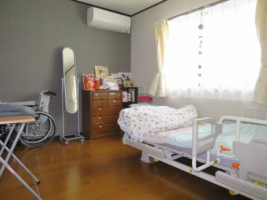 エアコン付きのフローリングの部屋には寝具付きのベッドが置かれ、チェストや車いす、鏡などもある。