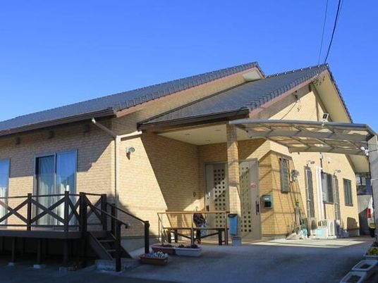 日当たりのいい場所に建っている平屋建ての建物はベージュの外壁で三角屋根が付き、ウッドデッキのテラスもある。