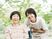 緑の多い屋外で笑顔で過ごしている車いすの高齢女性と付き添いのエプロンを付けた女性スタッフ。