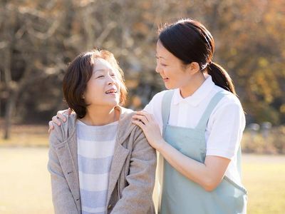 原っぱのような場所を笑顔で歩いている高齢女性と付き添いのブルーのエプロンを付けた若い女性スタッフ。