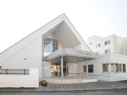 左側には手すり付きのスロープが設置された玄関があり、三角屋根が付いた建物がある。右側には白い大きな建物がある。