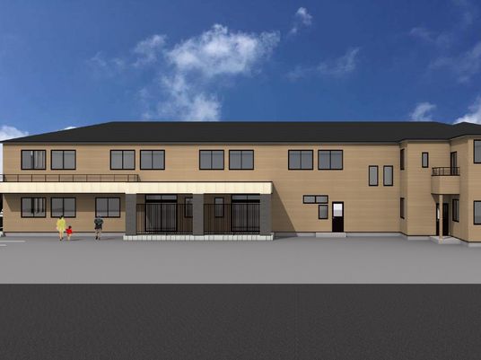 ベランダ付きの２階建ての建物はブラウン系で駐車場もあり、人が数名玄関に向かって歩いている。