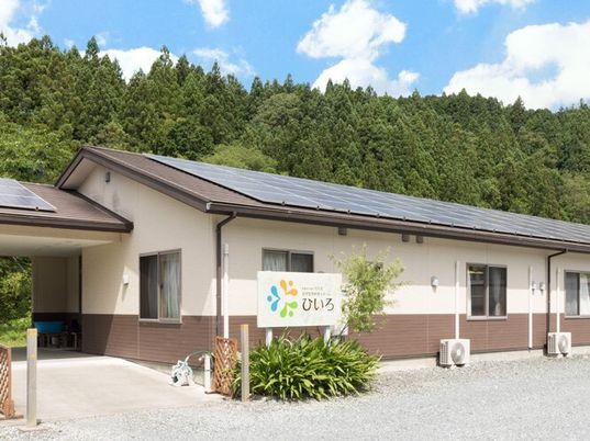 緑に囲まれた平屋建てのホーム。三角屋根が付き、玄関周りには緑が植えられ、ホーム名が書かれた看板もある。