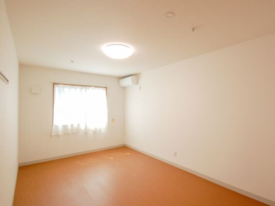 シンプルなフローリングの部屋にはエアコンや緊急通報装置があり、レースカーテンが付いた窓がある。