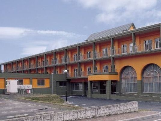 3階建ての建物。オレンジ系で各フロアにベランダが付き、駐車場もある。
