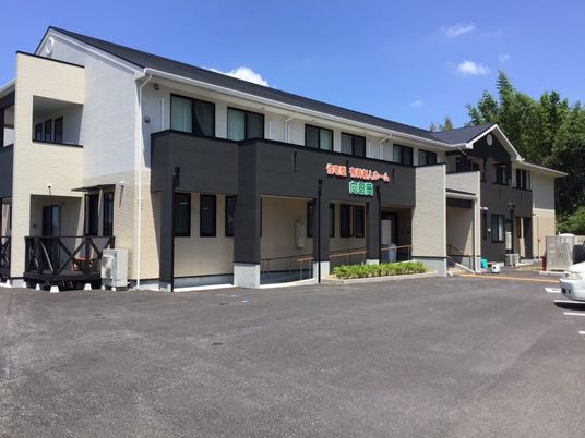 ベージュと黒の2階建ての建物。玄関先には手すり付きのスロープと階段があり、周りには緑が多くある。