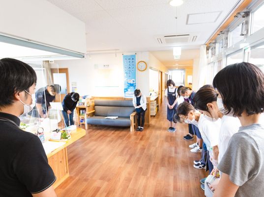 車いすの高齢女性にベージュのエプロンを付けた女性スタッフが付き添い、屋外での散策を楽しんでいる様子。